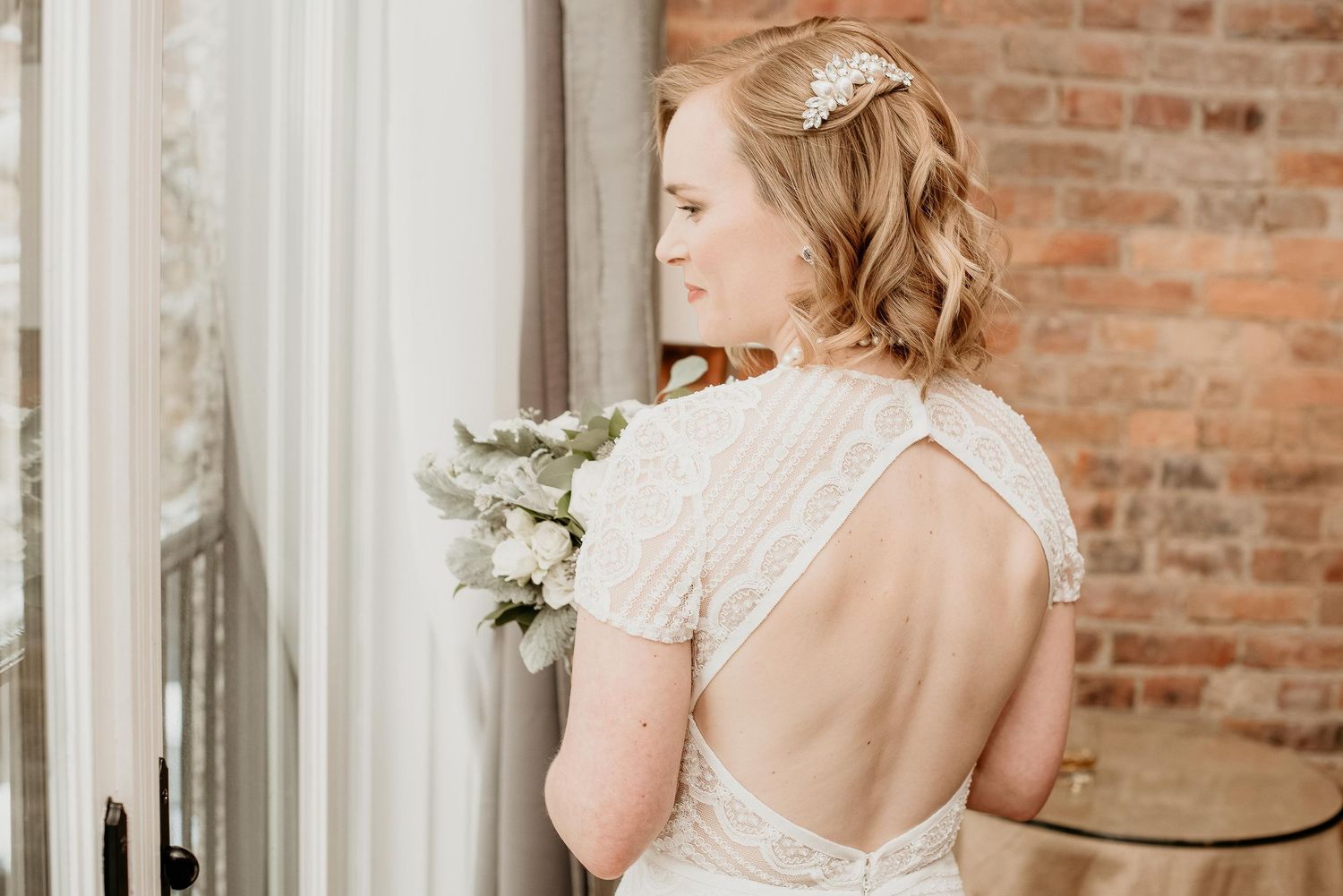 Bride in backless beaded white gown holding white bouquet, hair up with floral accessory, standing by window.