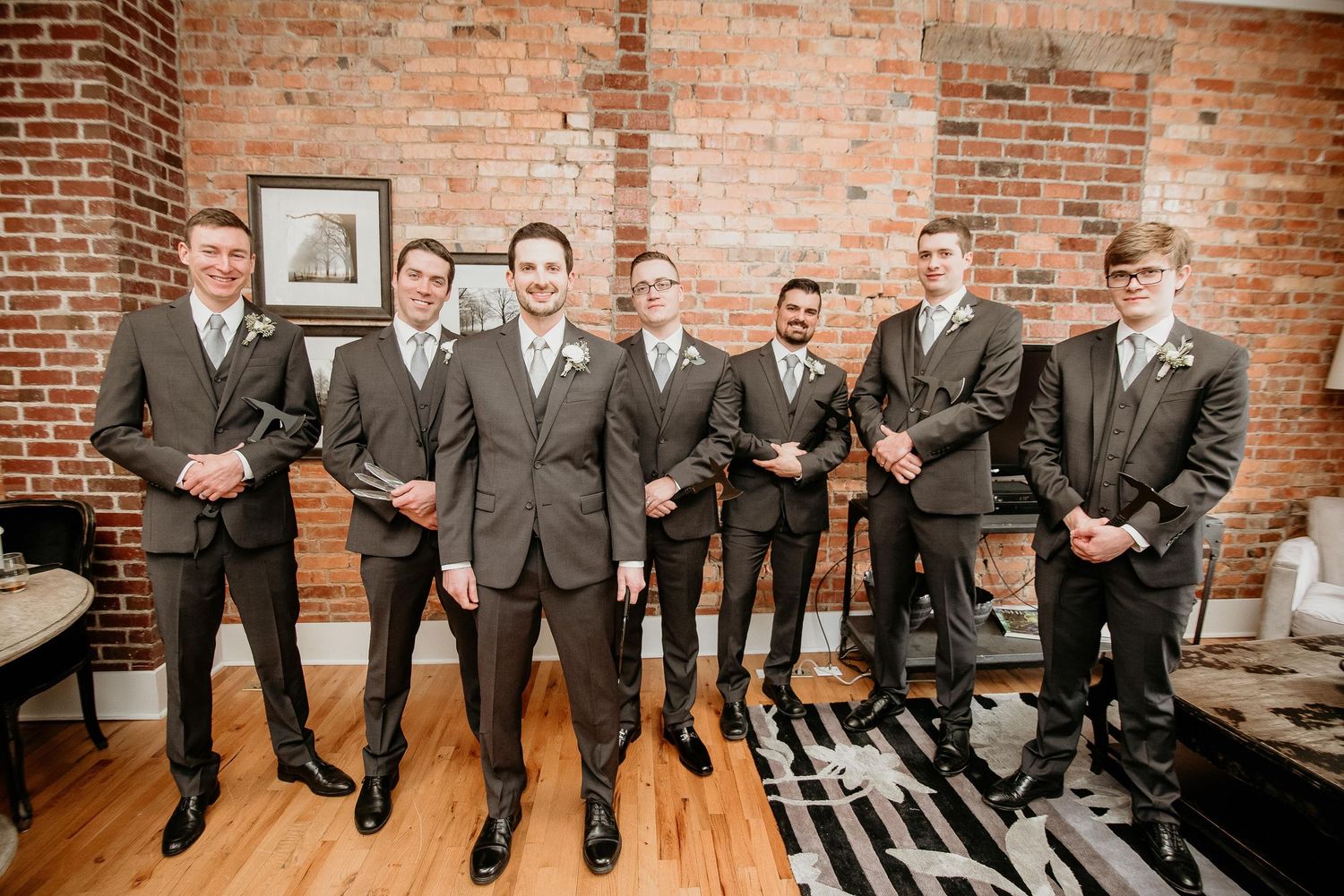 Groom and groomsmen in matching gray suits pose together in a rustic brick-walled venue with hardwood floors.