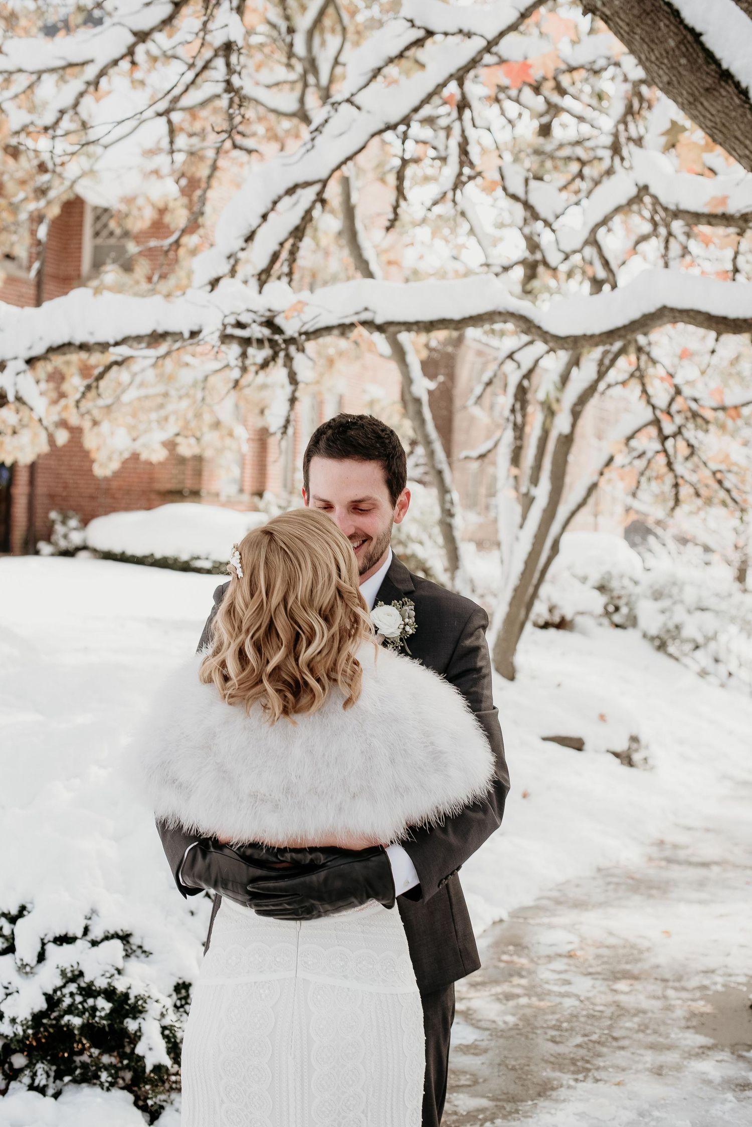 Couple embracing in snowy winter wedding, bride in white fur stole, groom in dark suit, snow-covered trees backdrop.