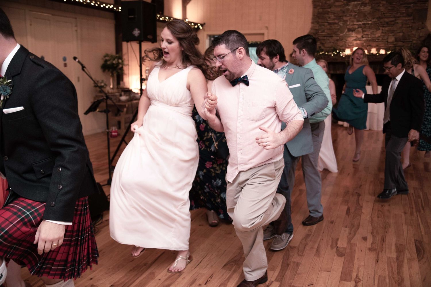Bride and groom dancing energetically at wedding reception on wooden dance floor with guests in background.