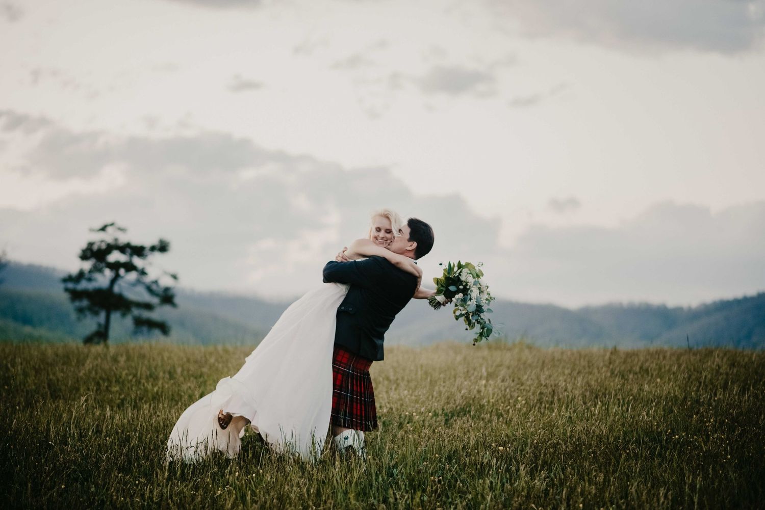 Groom in red tartan kilt lifts bride in white gown in lush green mountain meadow during romantic outdoor wedding.