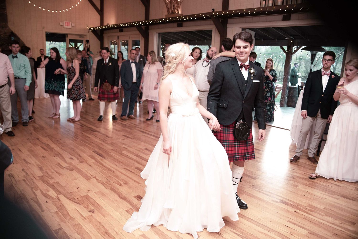 Bride in white gown and groom in red tartan kilt share first dance at rustic barn wedding reception.