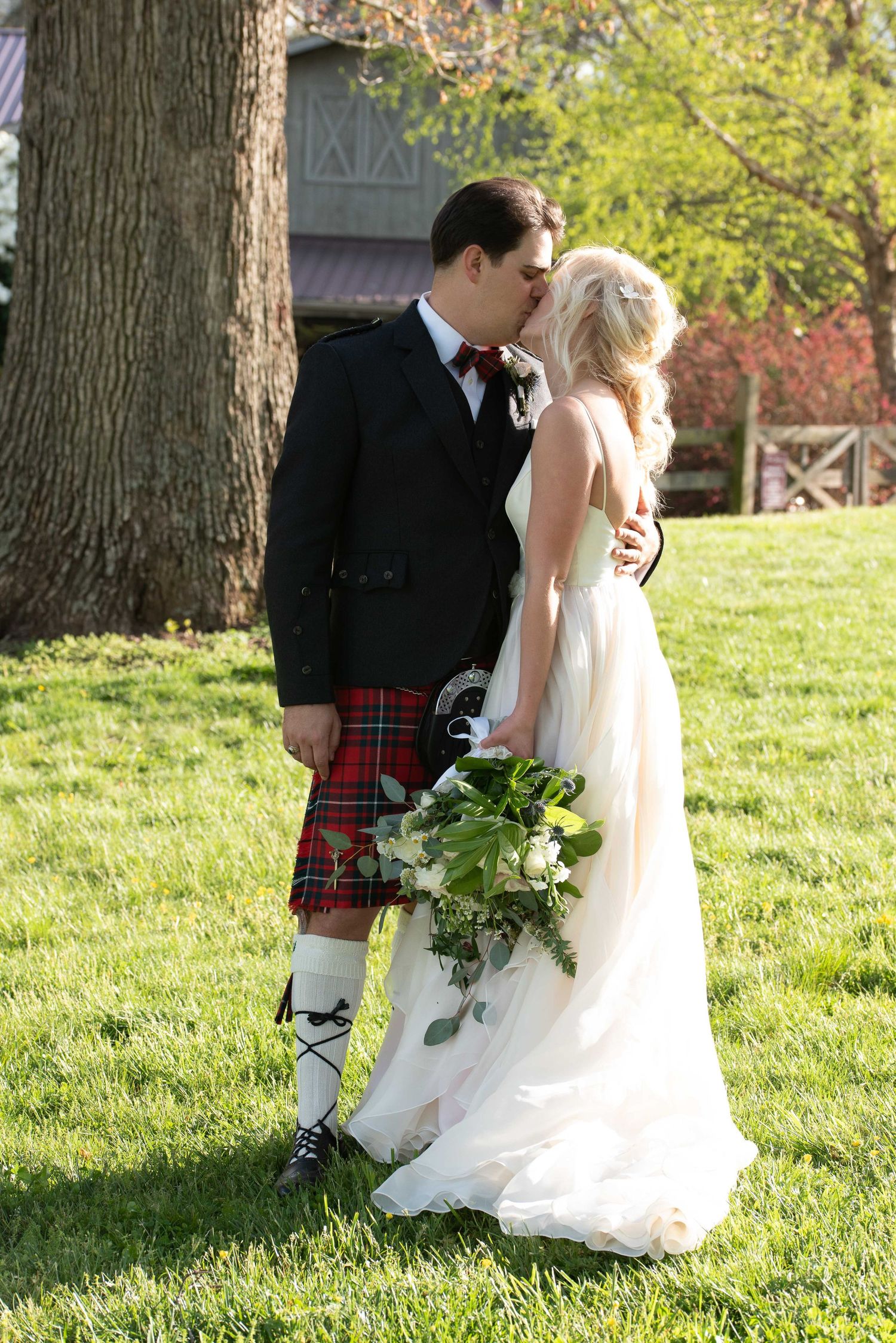 A bride and groom share a kiss outdoors, groom wearing a red tartan kilt and bow tie, bride holding a white bouquet.