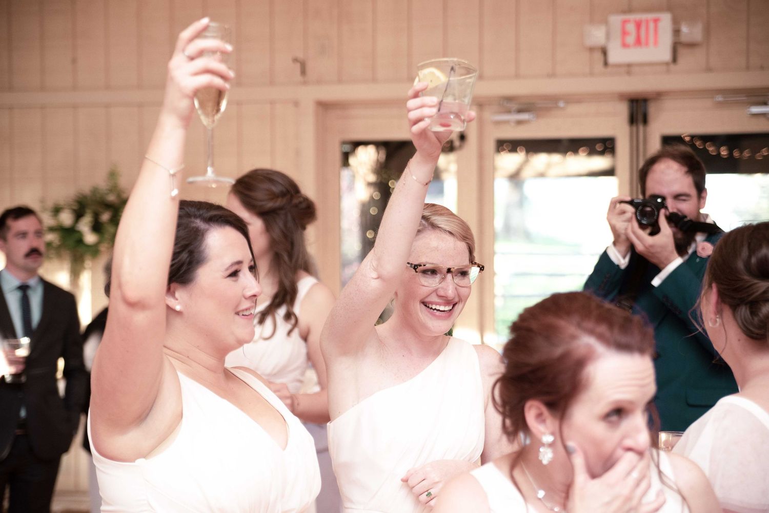 Two brides in white dresses raise champagne glasses in a joyful wedding toast surrounded by guests.