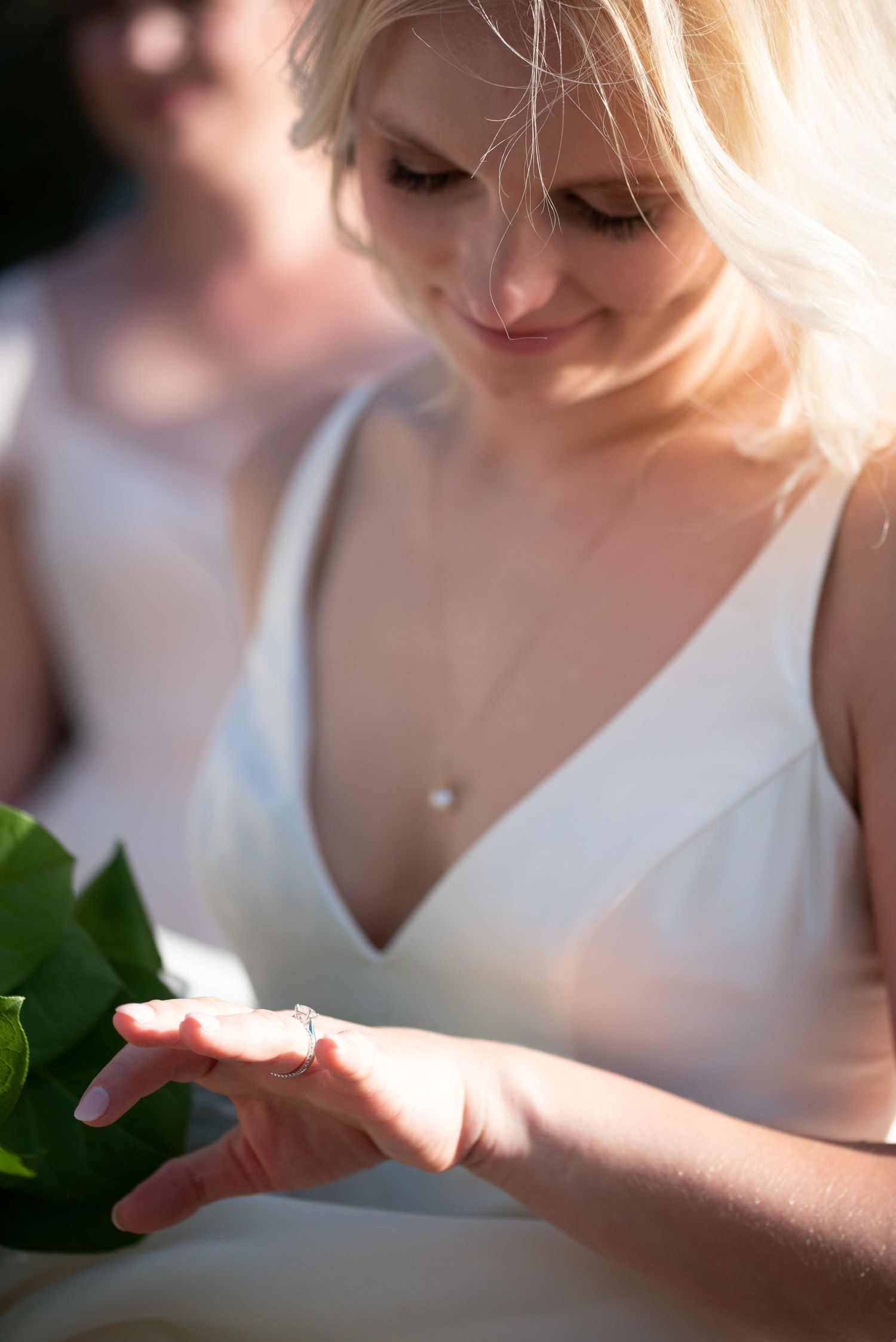 Blonde bride in white dress admiring her wedding ring while holding green leaves in sunlight.