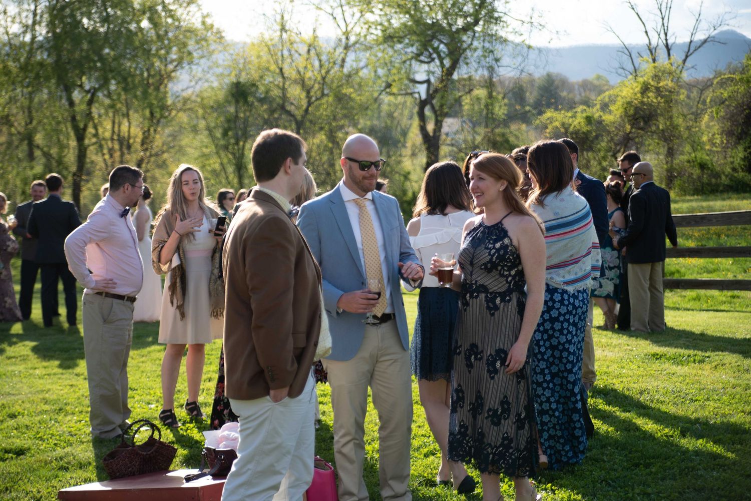 Guests mingling outdoors at a garden party or wedding reception on a sunny day with green hills in the background.