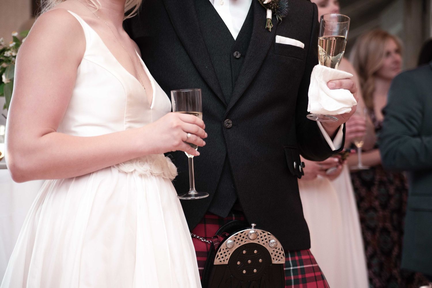 Bride and groom in Scottish kilt holding champagne glasses at wedding reception celebration.