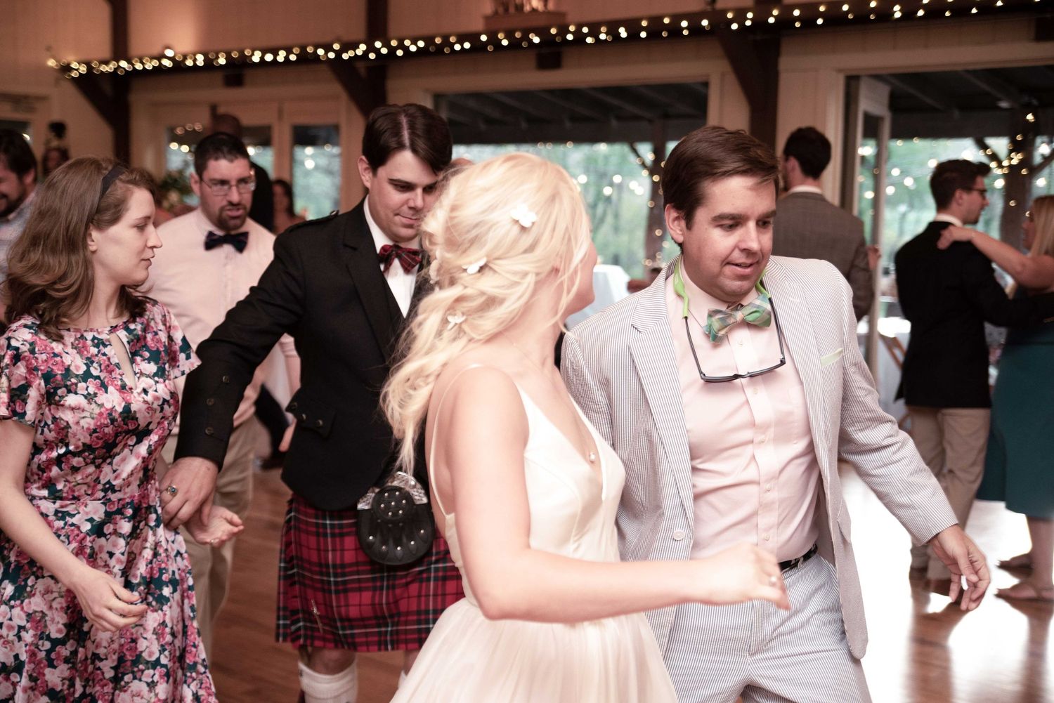 Bride and groom dancing joyfully at wedding reception with guests in formal attire including Scottish kilt in background.