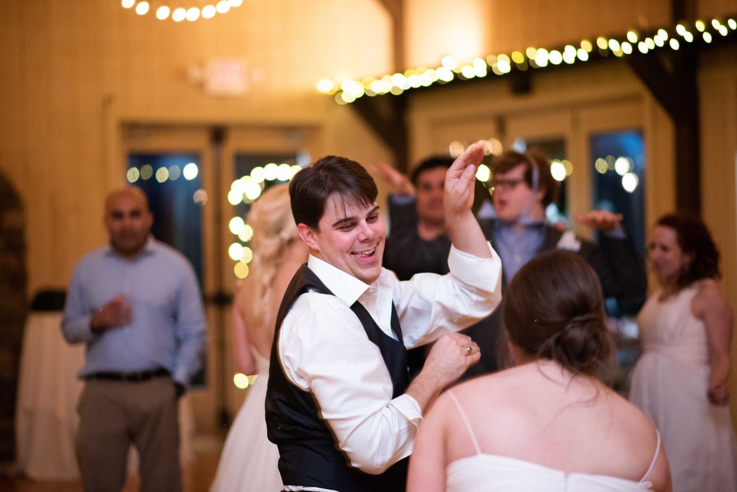 Groom dancing with bride at wedding reception, guests celebrating in background with warm string lights.