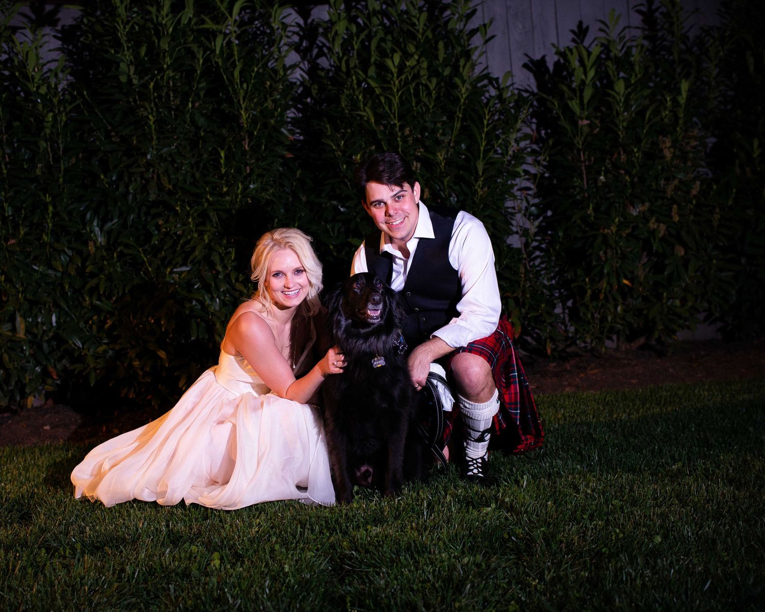 Bride and groom in Scottish kilt posing with black dog on green lawn at night wedding celebration.