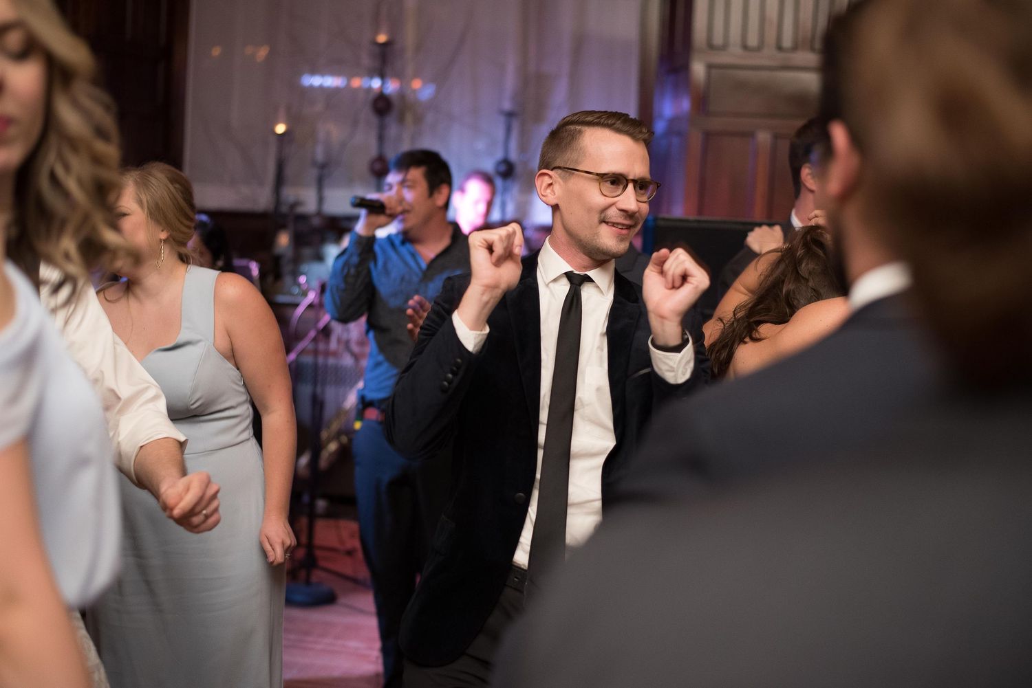 A man in a black suit dances joyfully at a wedding reception with guests and a live band in the background.