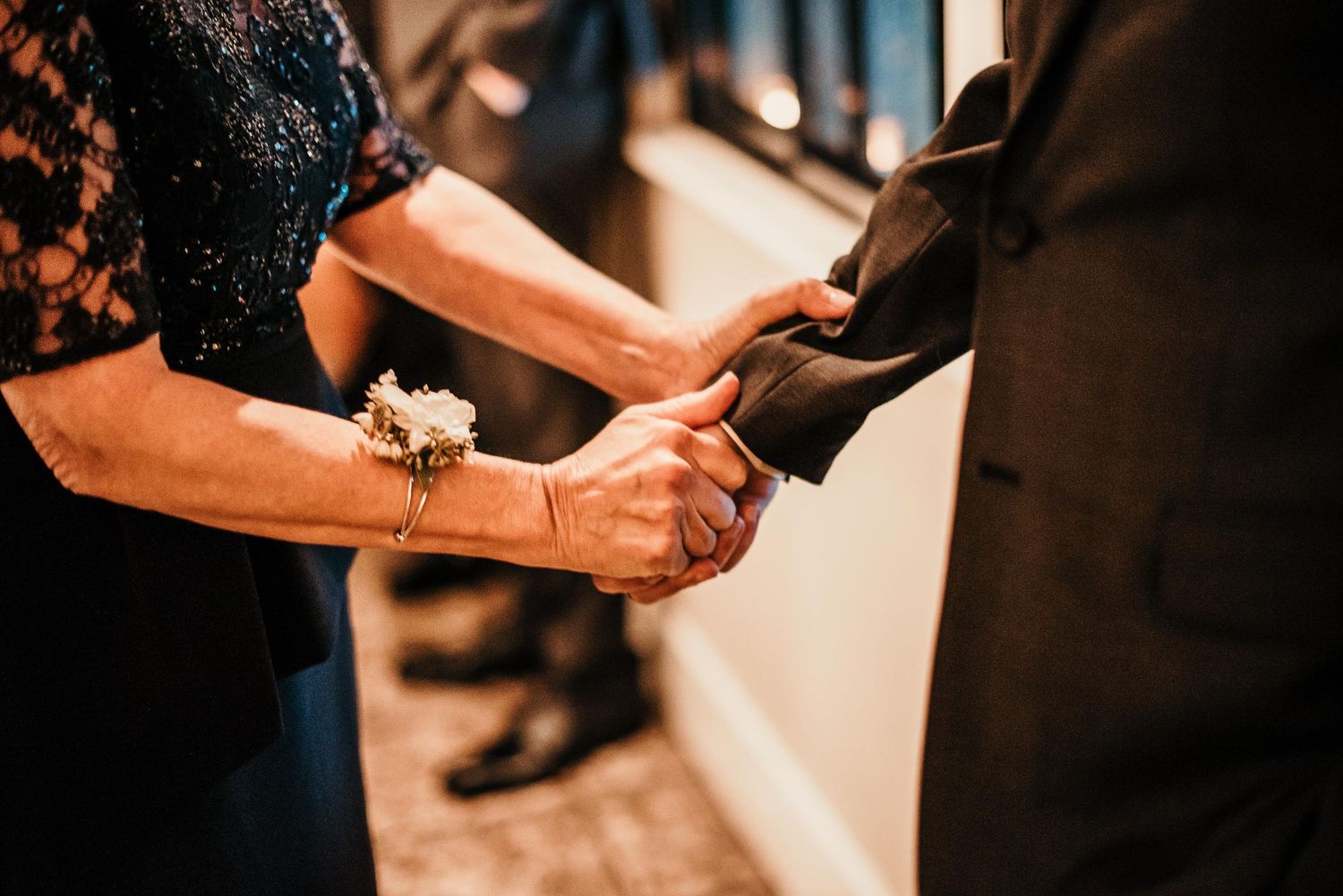 Close-up of two people holding hands at a formal event, woman wearing a floral wrist corsage and gold bracelet.