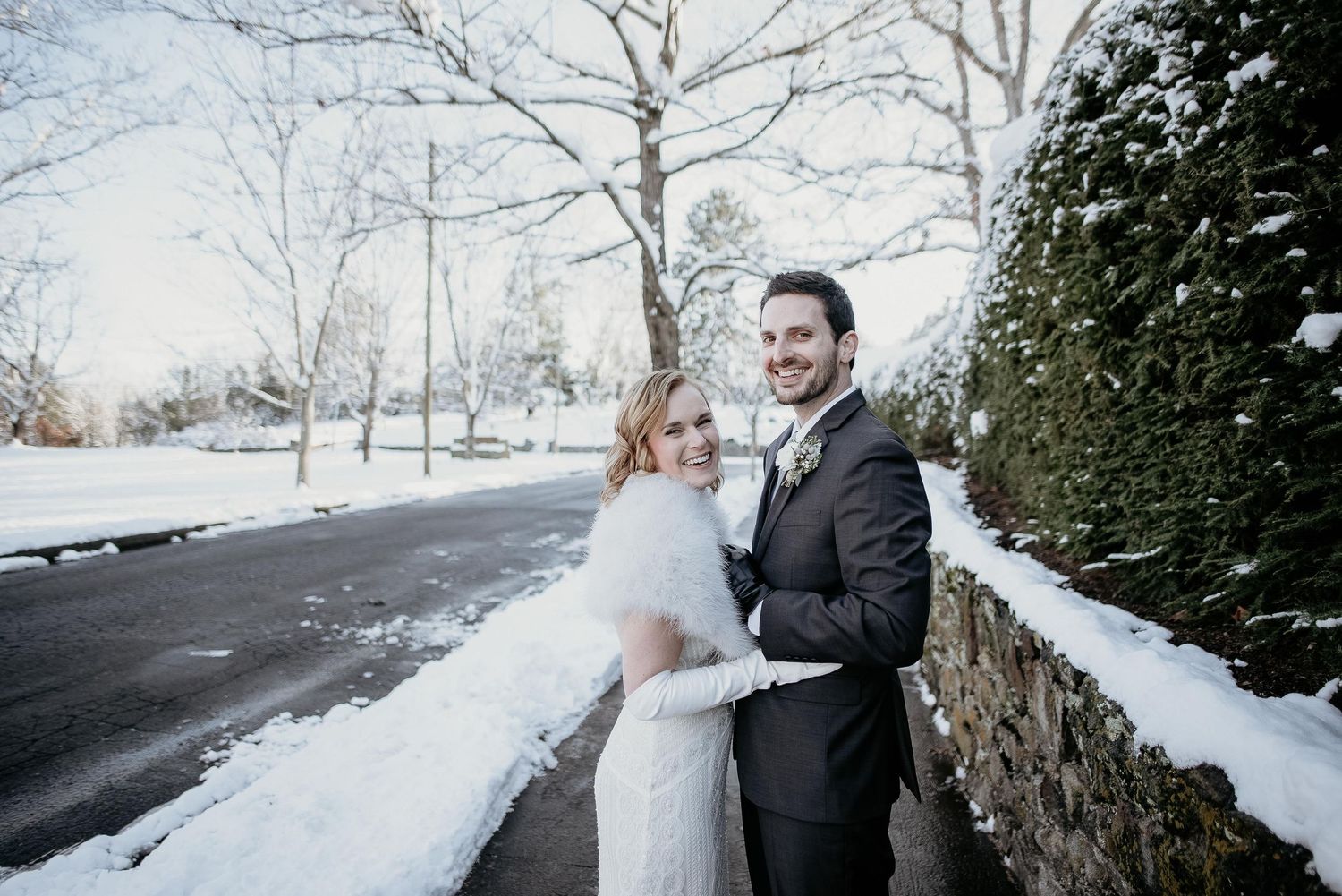 Bride and groom embrace outdoors in winter snow, she wears a white fur wrap and he wears a dark suit.