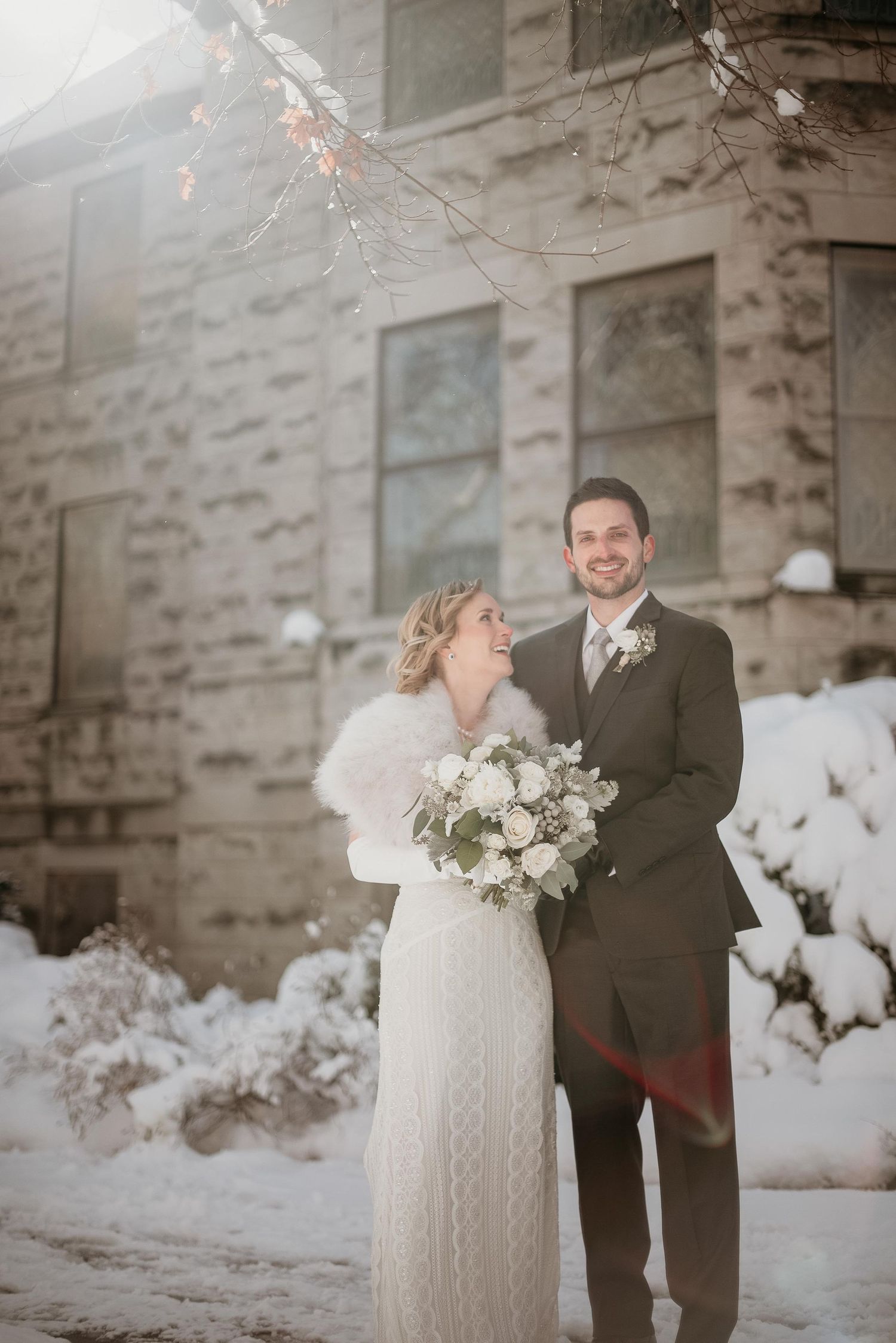 Bride and groom pose together in snow outside stone building, bride holding white bouquet wearing fur wrap over wedding dress.