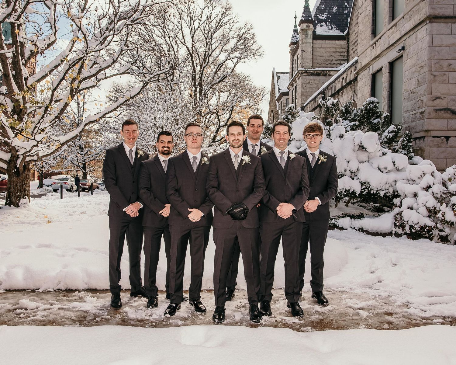 Groomsmen in dark suits pose outdoors in snowy winter setting near historic building for wedding photo.