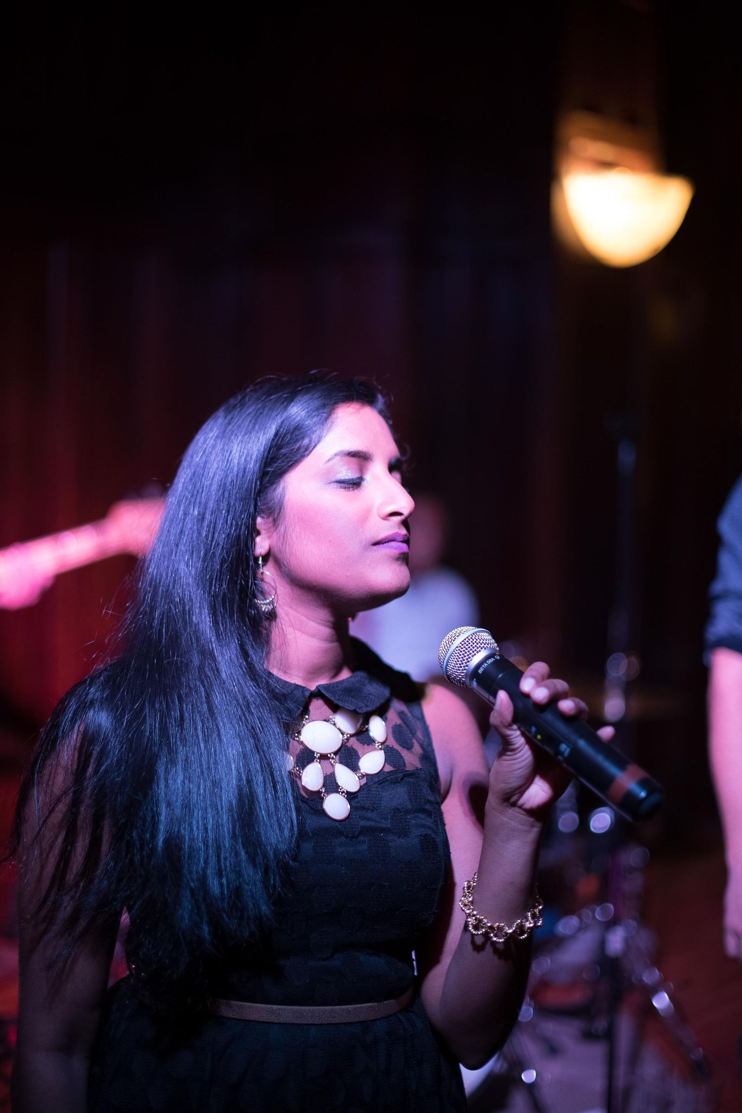 Young woman with long dark hair singing into a microphone on stage, wearing a black dress under warm stage lights.