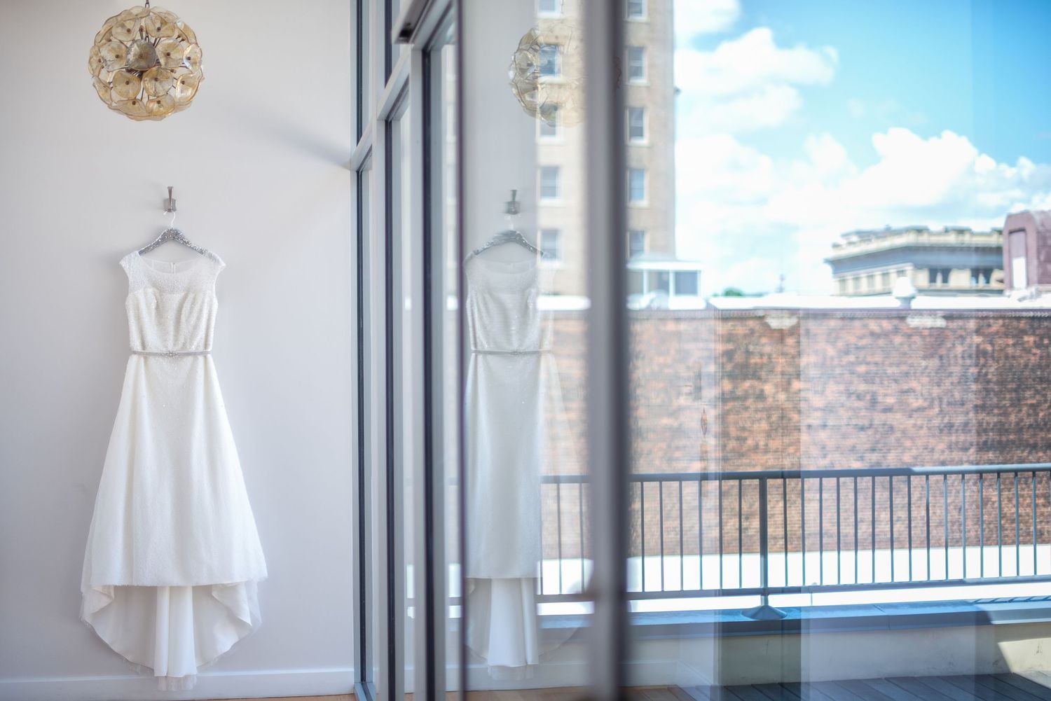 White wedding dress hanging on wall beside floor-to-ceiling windows with city skyline view on sunny day.