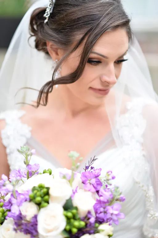 Bride in white lace gown holding purple and white floral bouquet, wearing veil with hair up.