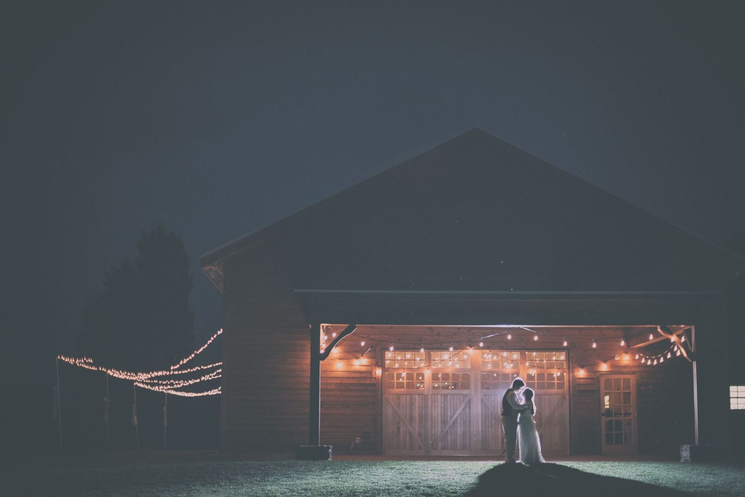 Romantic couple silhouetted against a rustic barn adorned with warm string lights at night.