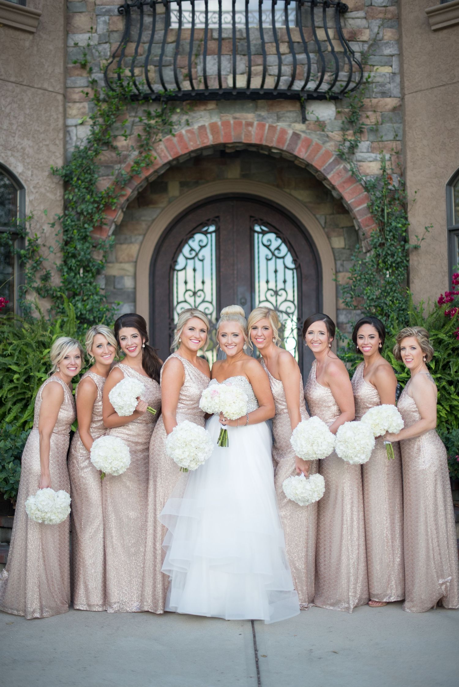 Bride with bridesmaids in champagne sequin gowns holding white hydrangea bouquets outside elegant venue.