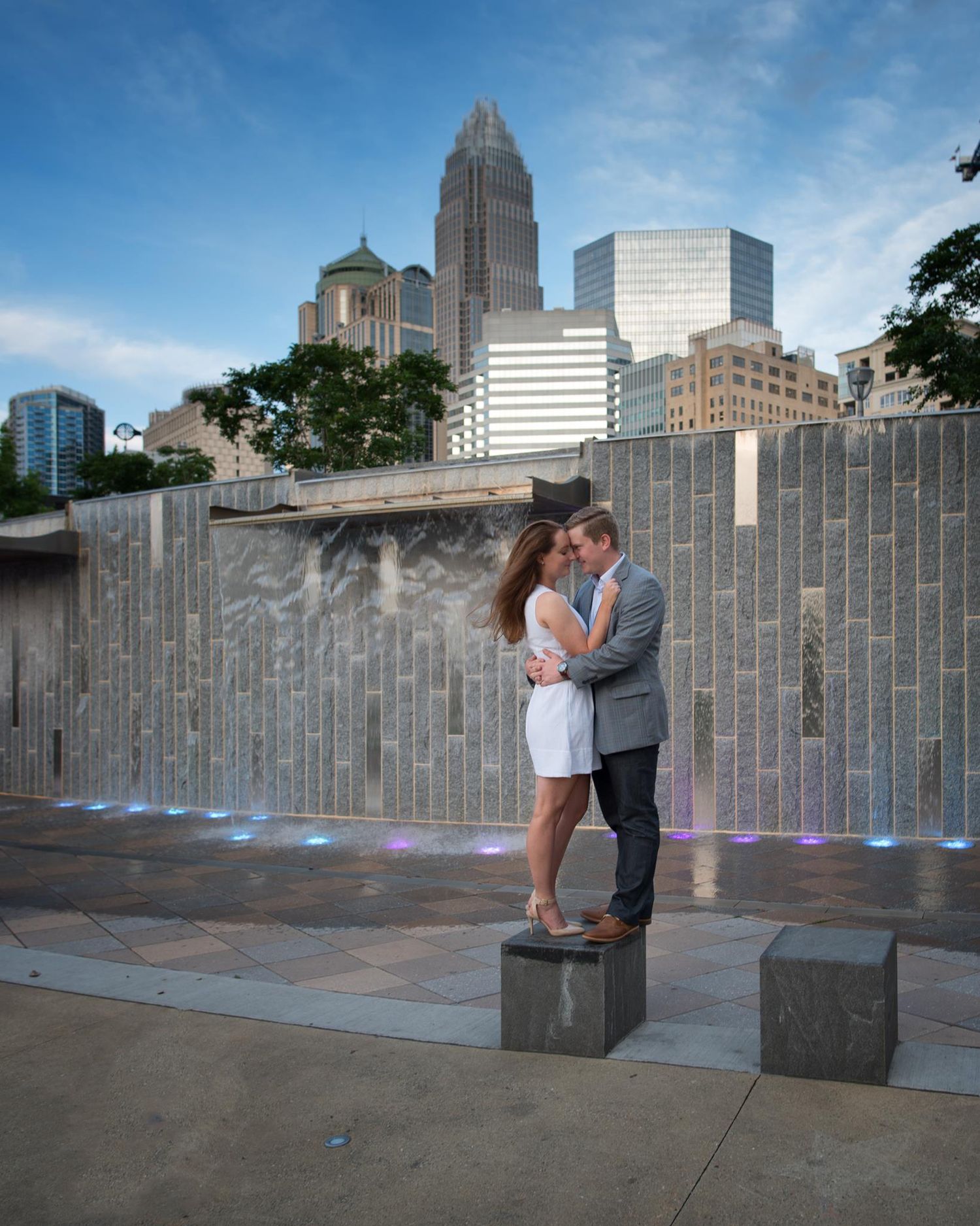 Couple embracing romantically in front of a waterfall wall with Charlotte city skyline.