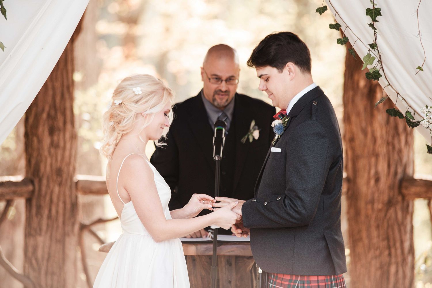 Bride and groom exchange rings at outdoor wedding ceremony with officiant, groom wearing traditional Scottish kilt.