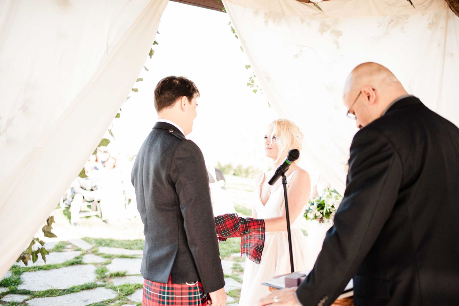 Outdoor wedding ceremony with groom in tartan kilt, blonde officiant holding microphone, and guest under white draped canopy.