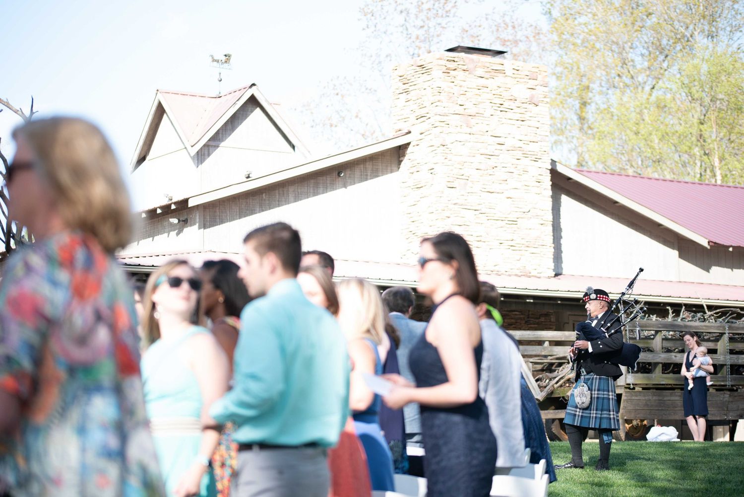 Wedding guests gather outdoors near a rustic venue as a bagpiper performs in traditional attire.