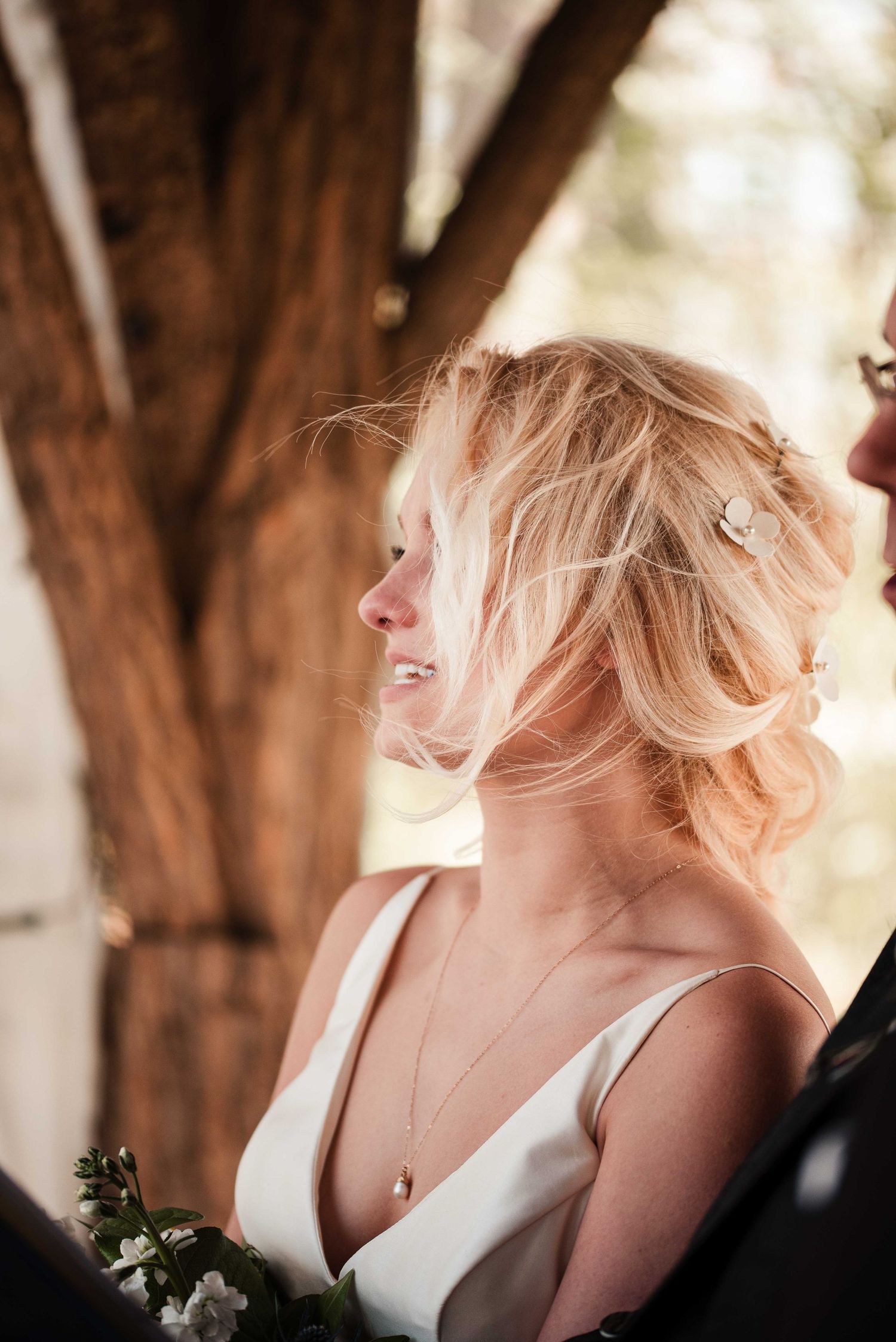 Blonde bride in white gown with floral hair pin holds bouquet outdoors near rustic tree backdrop.