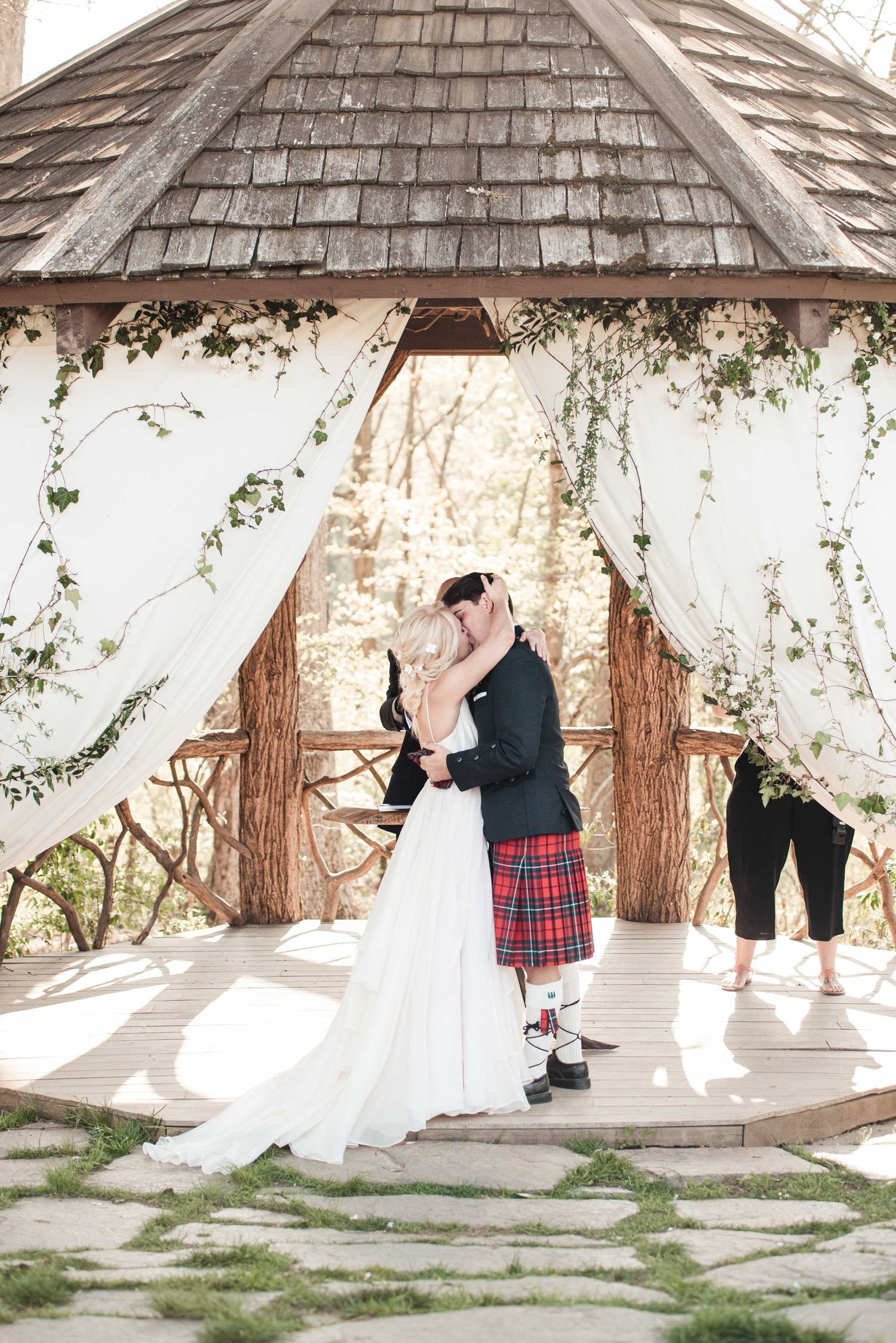 Bride and groom share first kiss under rustic wooden gazebo with white draping and ivy at outdoor wedding ceremony.