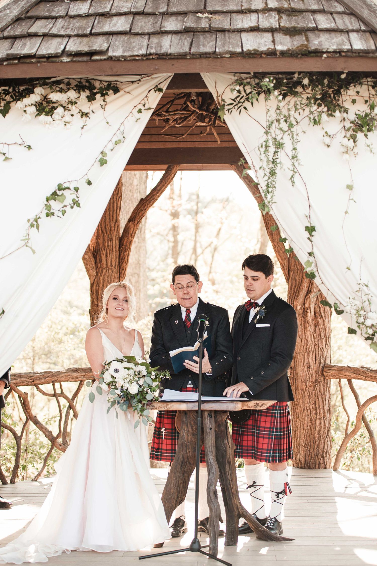 Bride and groom exchange vows under a rustic gazebo adorned with white drapes and greenery, officiant in a tartan kilt.