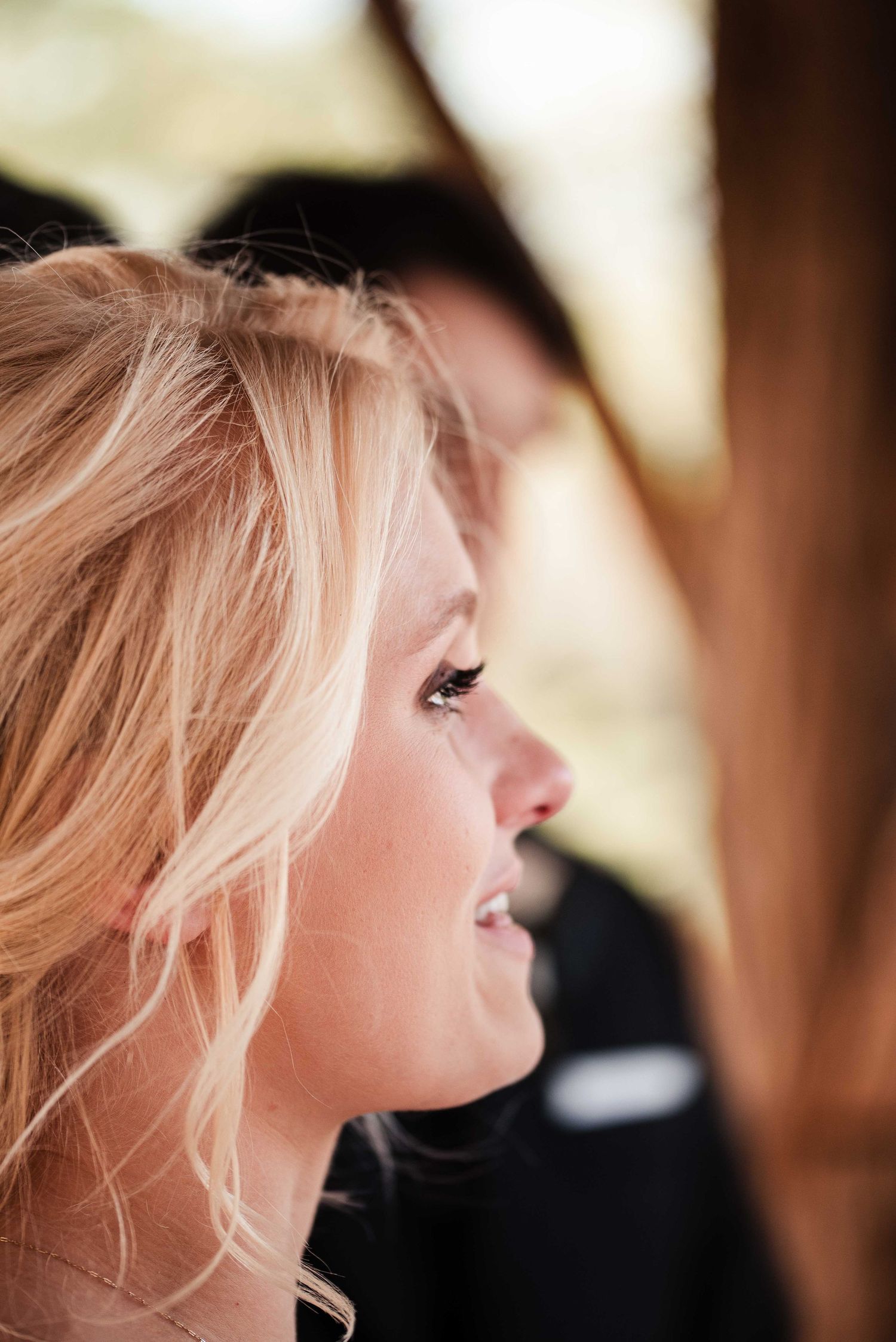 Close-up profile of a blonde woman with wavy hair gazing upward, with a blurred figure in dark clothing behind her.