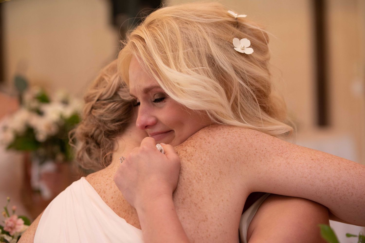Two women in white dresses share an emotional hug at a wedding ceremony, one with blonde hair adorned with a flower.