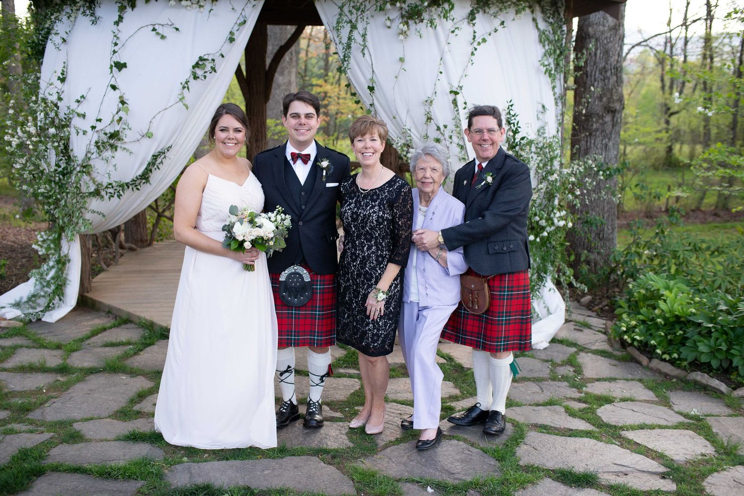 Wedding party posing under floral arch, bride in white gown with two men in red kilts and woman in black dress.