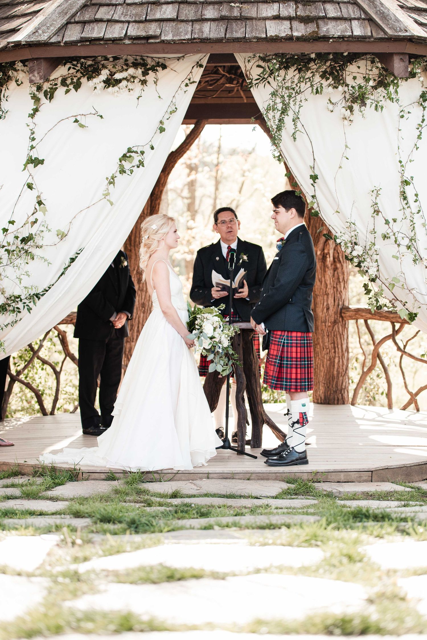 Outdoor wedding ceremony under rustic wooden gazebo with draped fabric, bride in white gown, groom in blue jacket and red kilt.