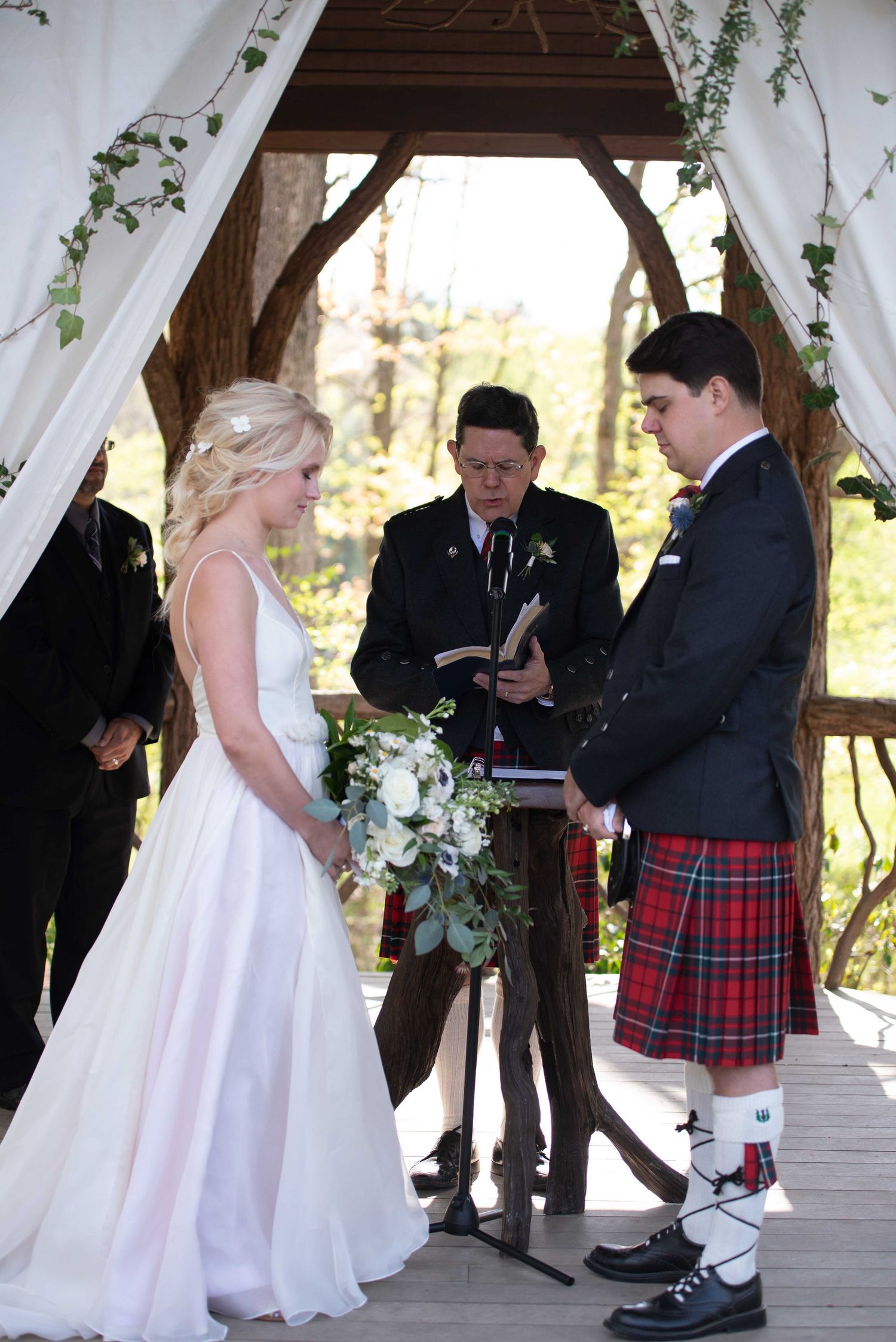 Bride in white gown and groom in red tartan kilt exchange vows at outdoor woodland wedding ceremony.
