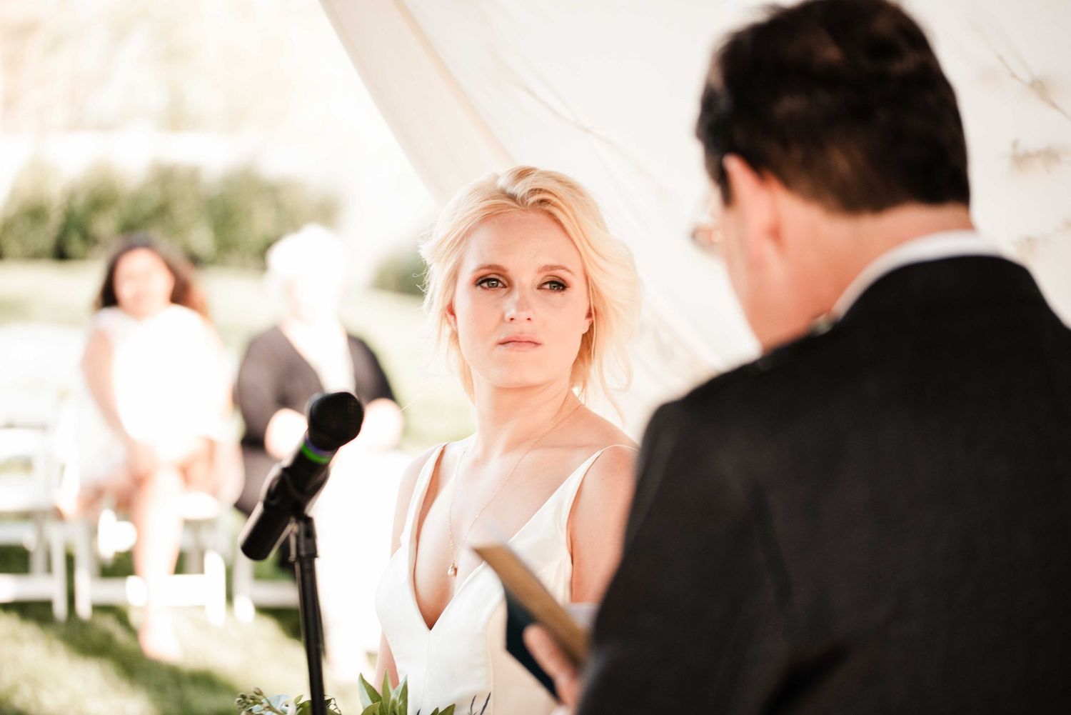 Blonde bride in white gown gazes at groom during outdoor wedding ceremony under elegant tent.
