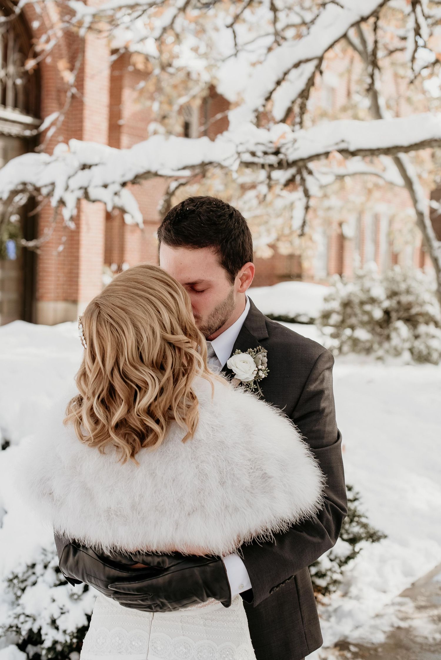 Bride and groom sharing a kiss in a snowy winter wonderland, bride wearing a white fur wrap and groom in a dark suit.