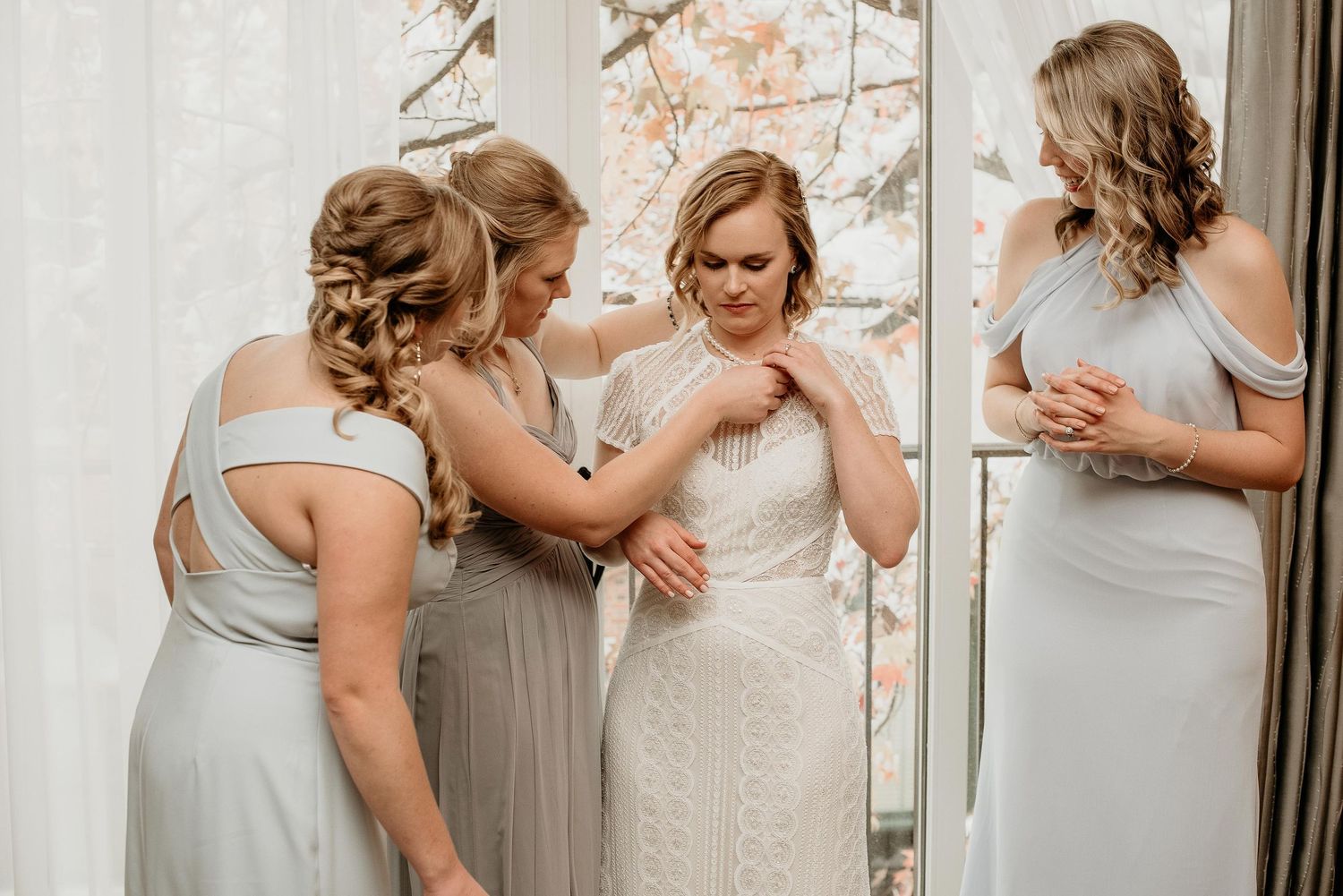 Bridesmaids helping bride into her lace wedding dress near a mirror and window on her wedding day.