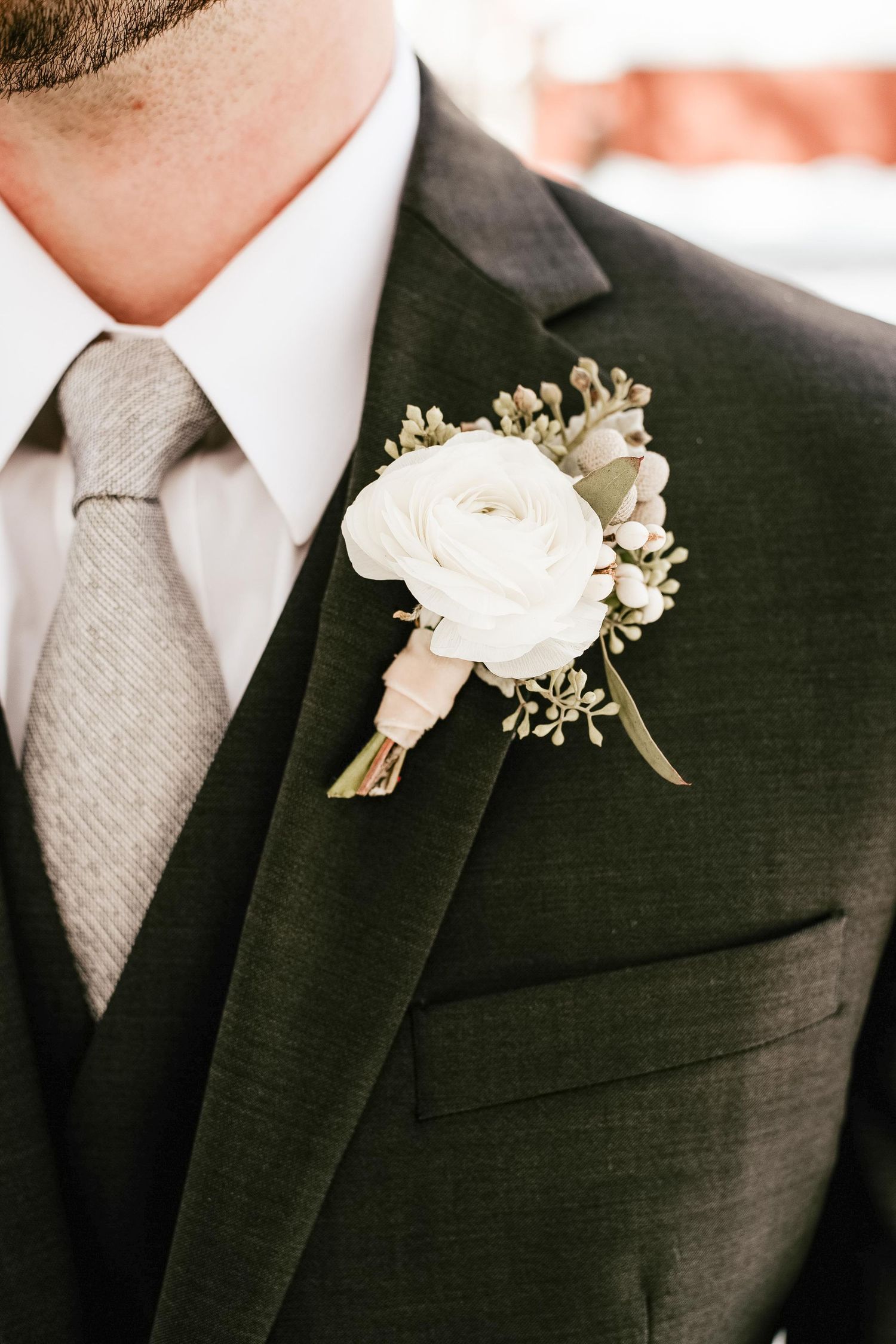Groom in dark green suit with white ranunculus boutonniere, gray tie, and white dress shirt at wedding.