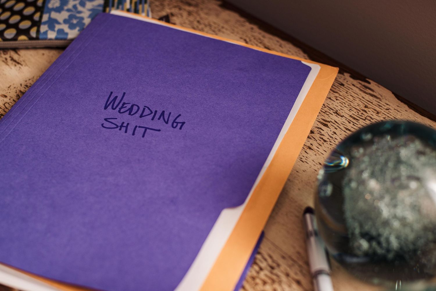 Purple wedding planner notebook labeled 'Wedding Shit' in black marker, with a pen and decorative glass orb nearby.
