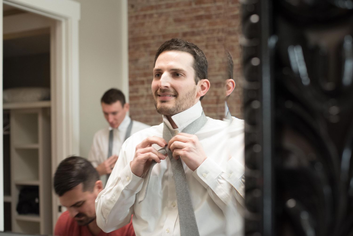 Groom adjusting his gray tie while getting ready for wedding ceremony in a brick-walled room.