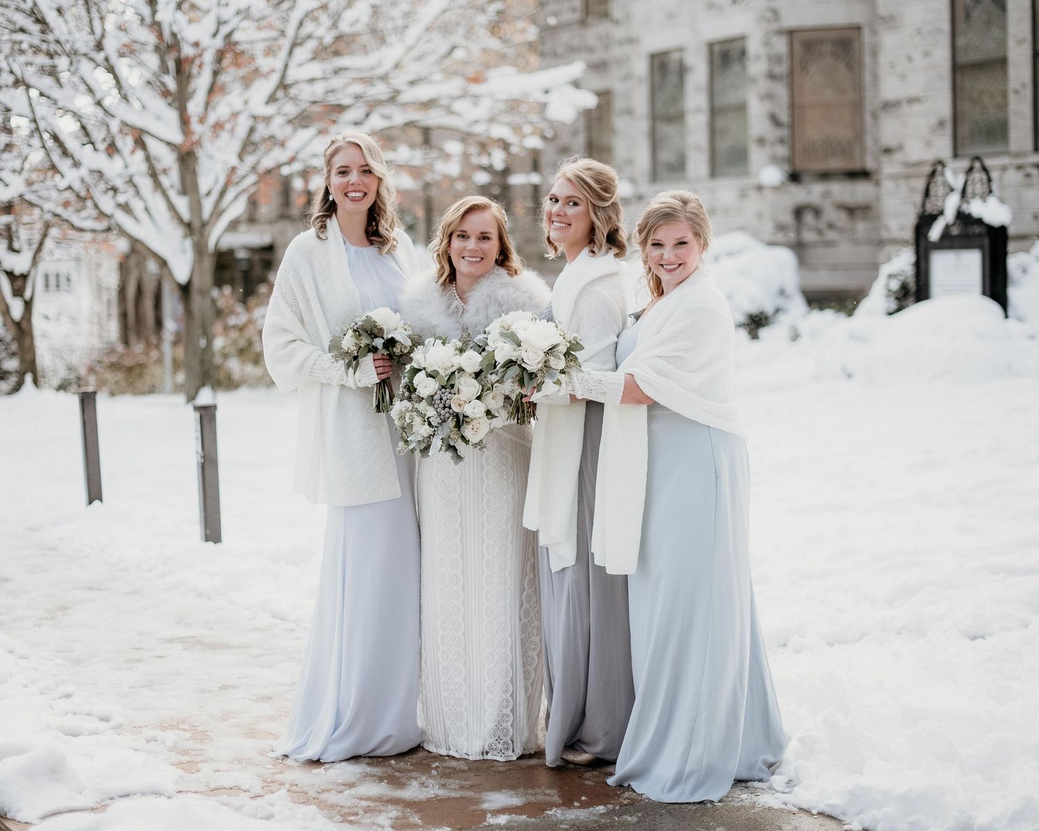 Bride and bridesmaids in white wraps and light blue gowns holding bouquets outdoors in a snowy winter wedding setting.