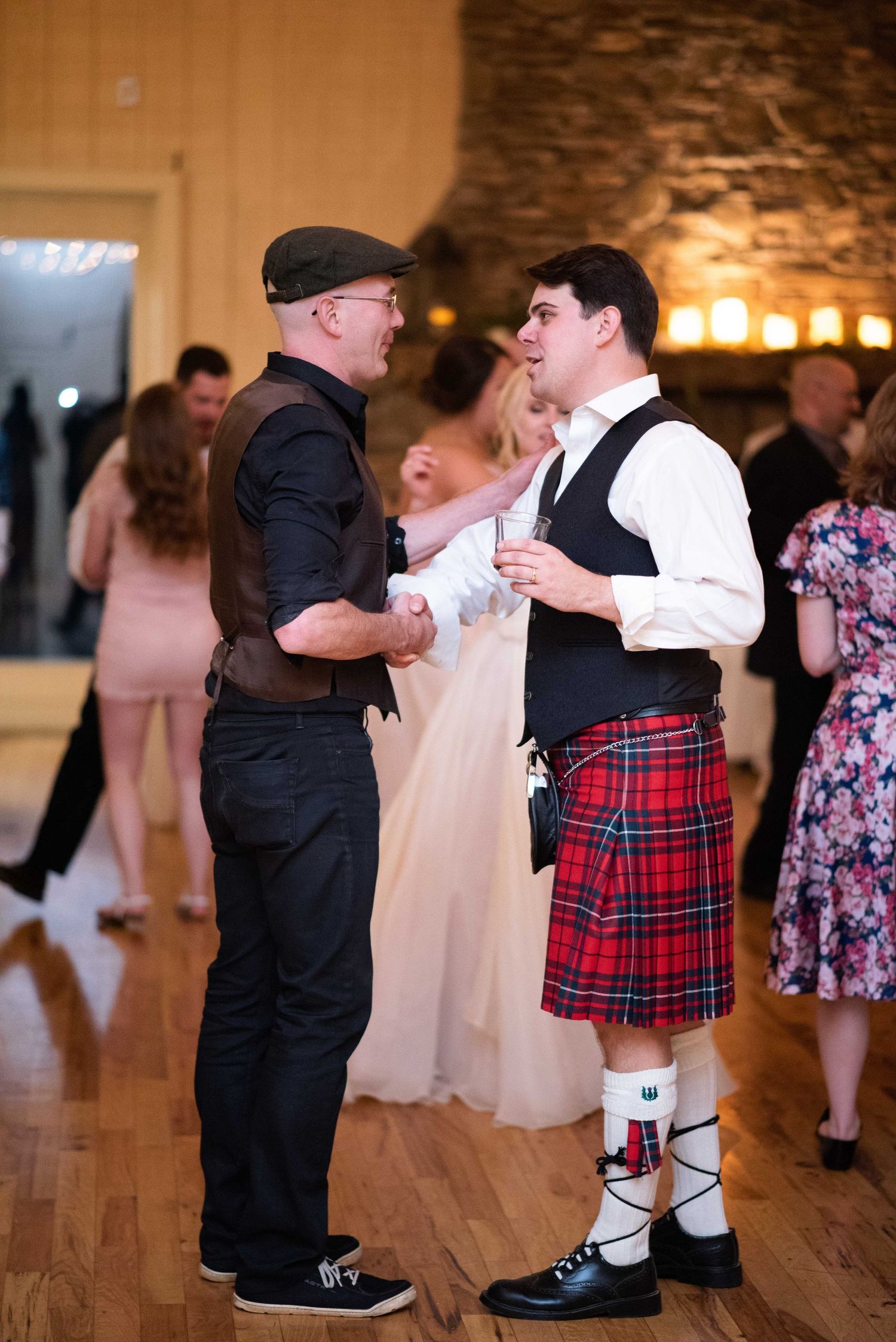 Two men converse at a wedding reception, one wearing a traditional red tartan kilt with white knee socks and ghillie brogues.
