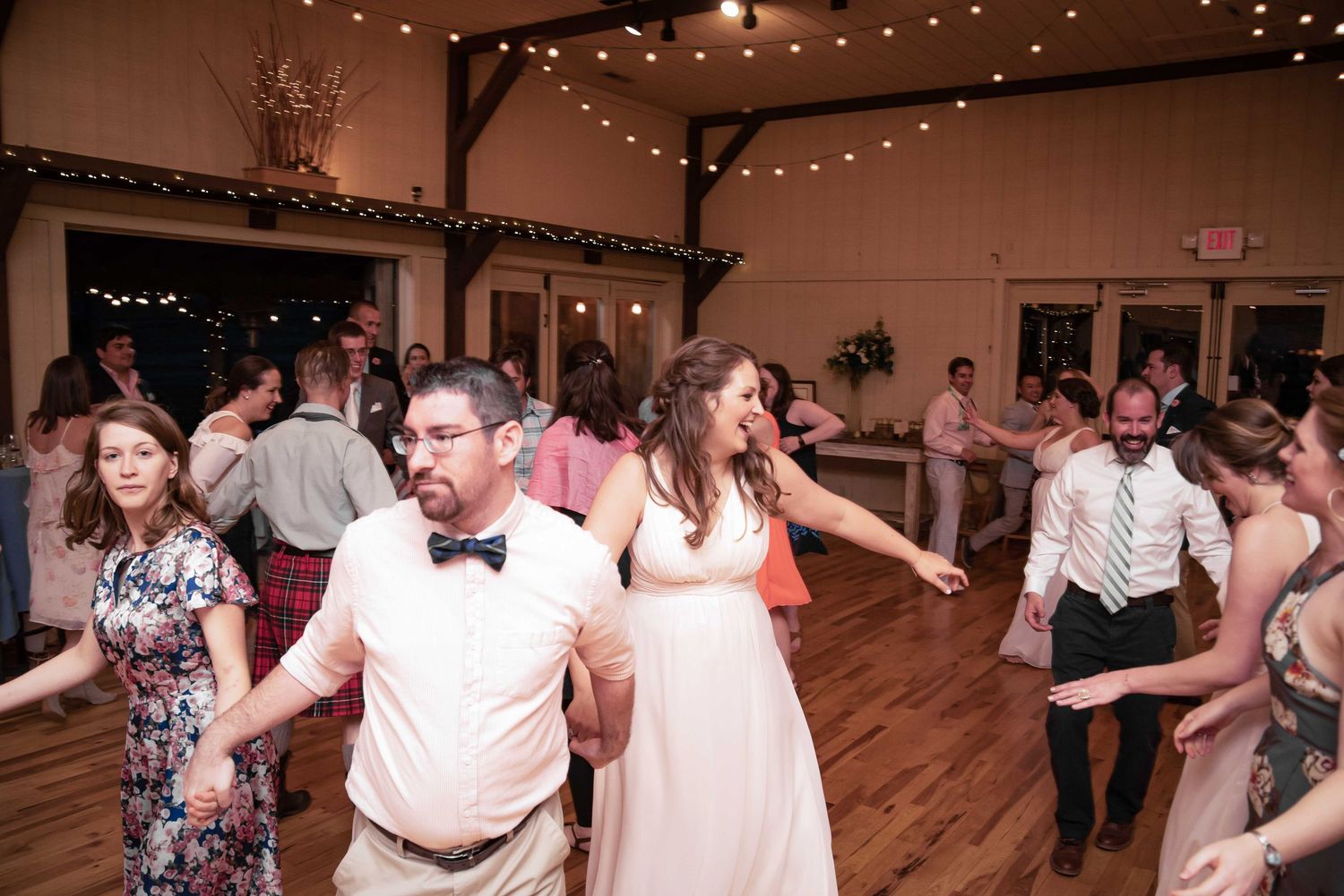 Bride and groom dancing joyfully with guests at a rustic wedding reception venue with string lights.