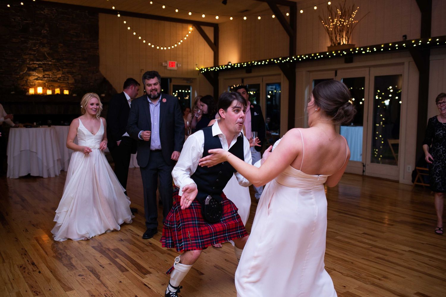 Wedding guests dancing at a rustic venue, featuring a person in a red tartan kilt during a lively reception celebration.
