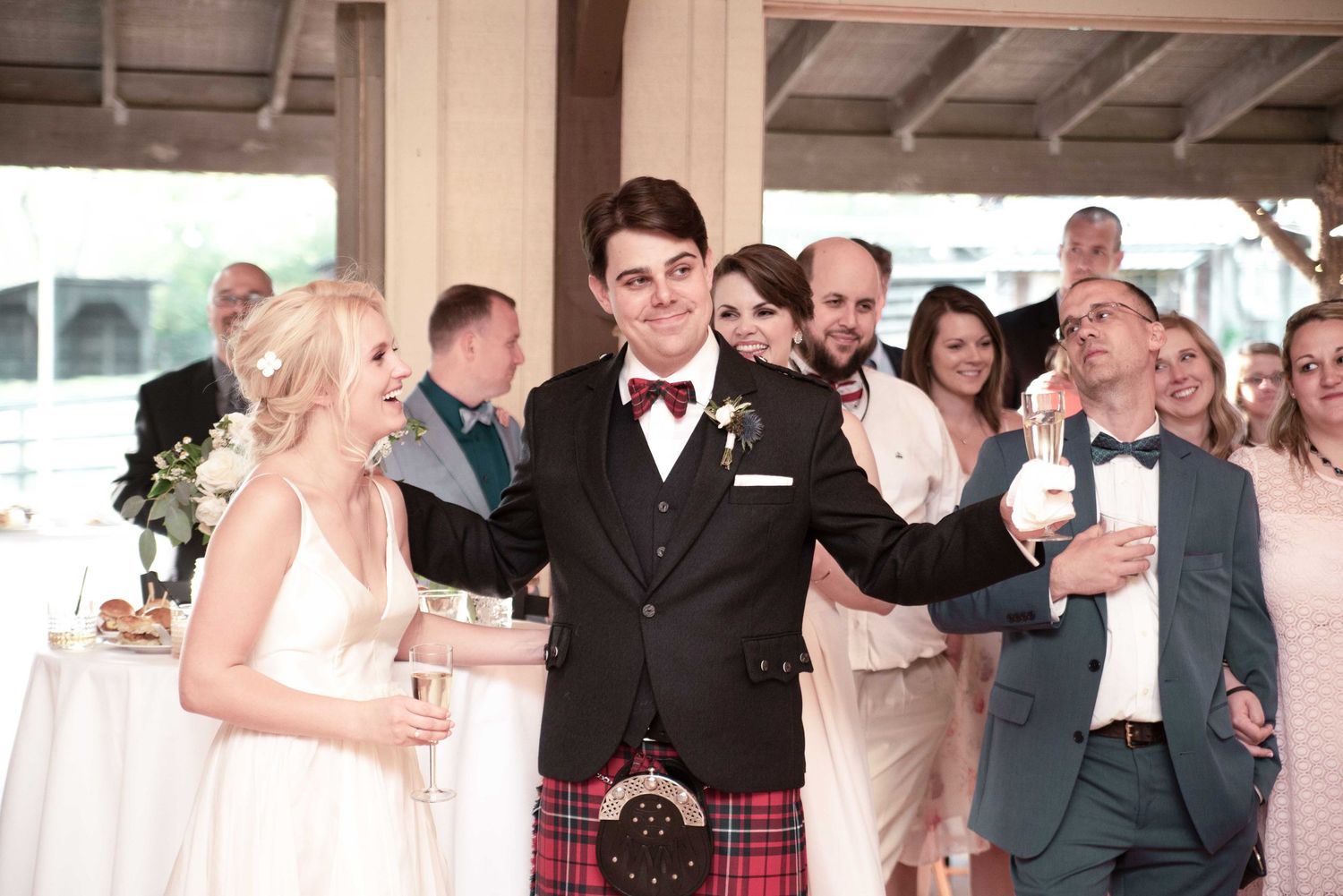 Groom in tartan kilt and bow tie raises a toast with bride in white dress at elegant wedding reception.