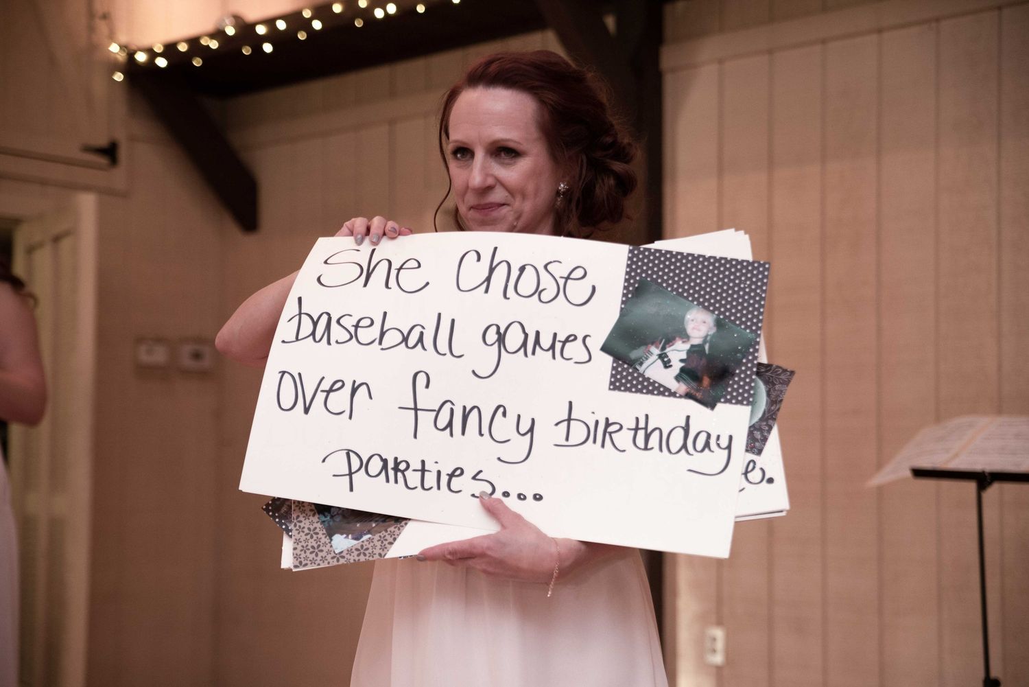 Woman holding a handwritten sign reading she chose baseball games over fancy birthday parties at a wedding reception.