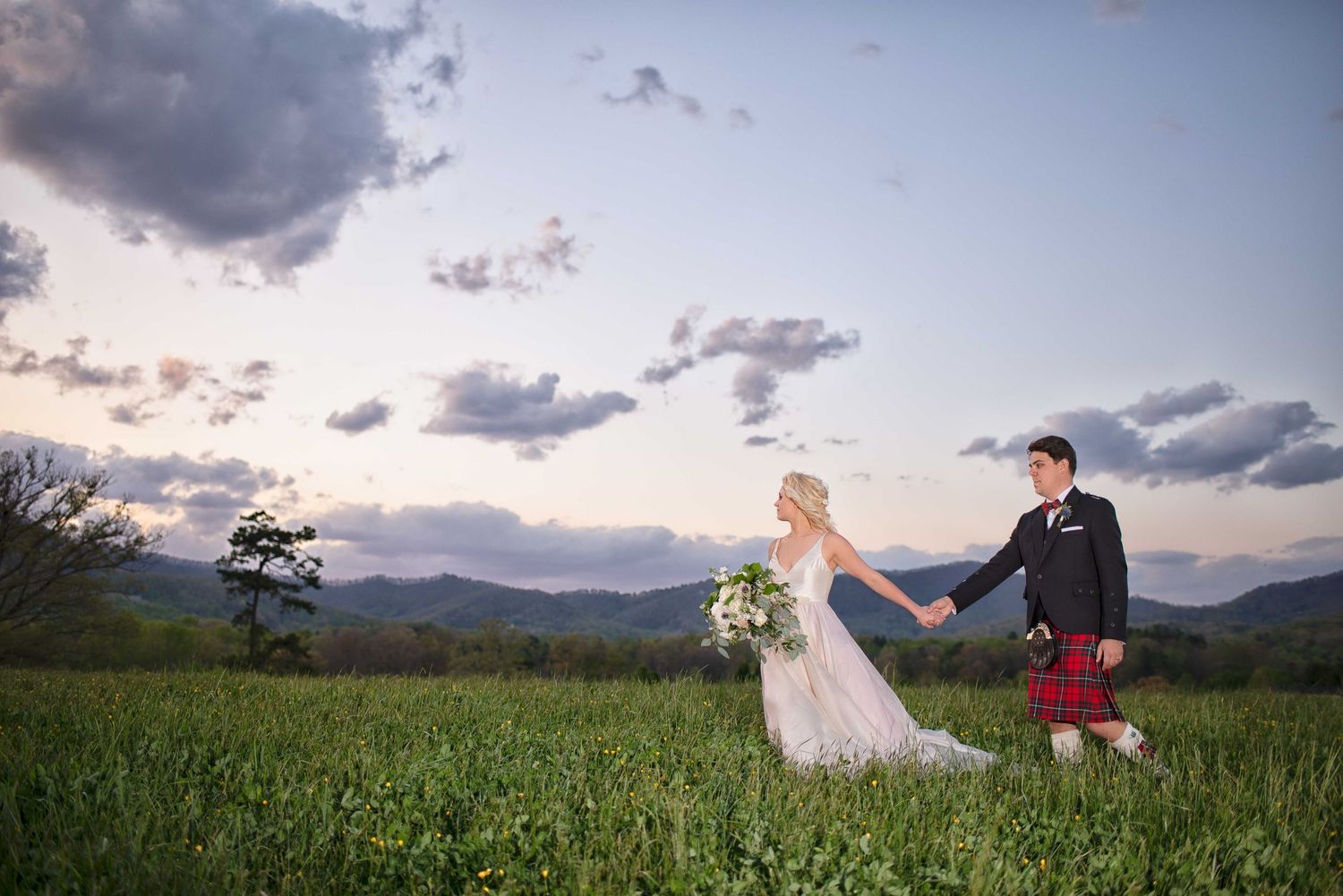 Bride and groom hold hands in a lush green field at sunset with misty mountains in the background.