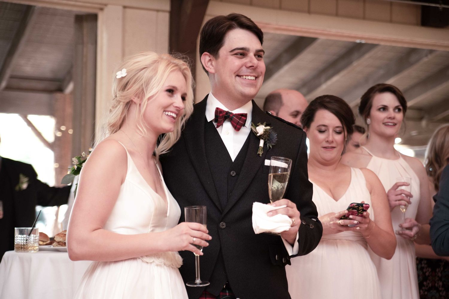 Bride and groom smiling and holding champagne glasses at their wedding reception with bridesmaids in background.
